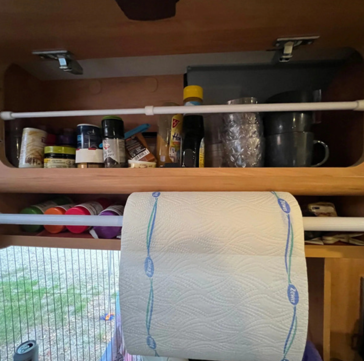 Retenue anti-chute grand modèle installée devant les provisions d’un placard, avec une seconde barre portant un rouleau d’essuie-tout